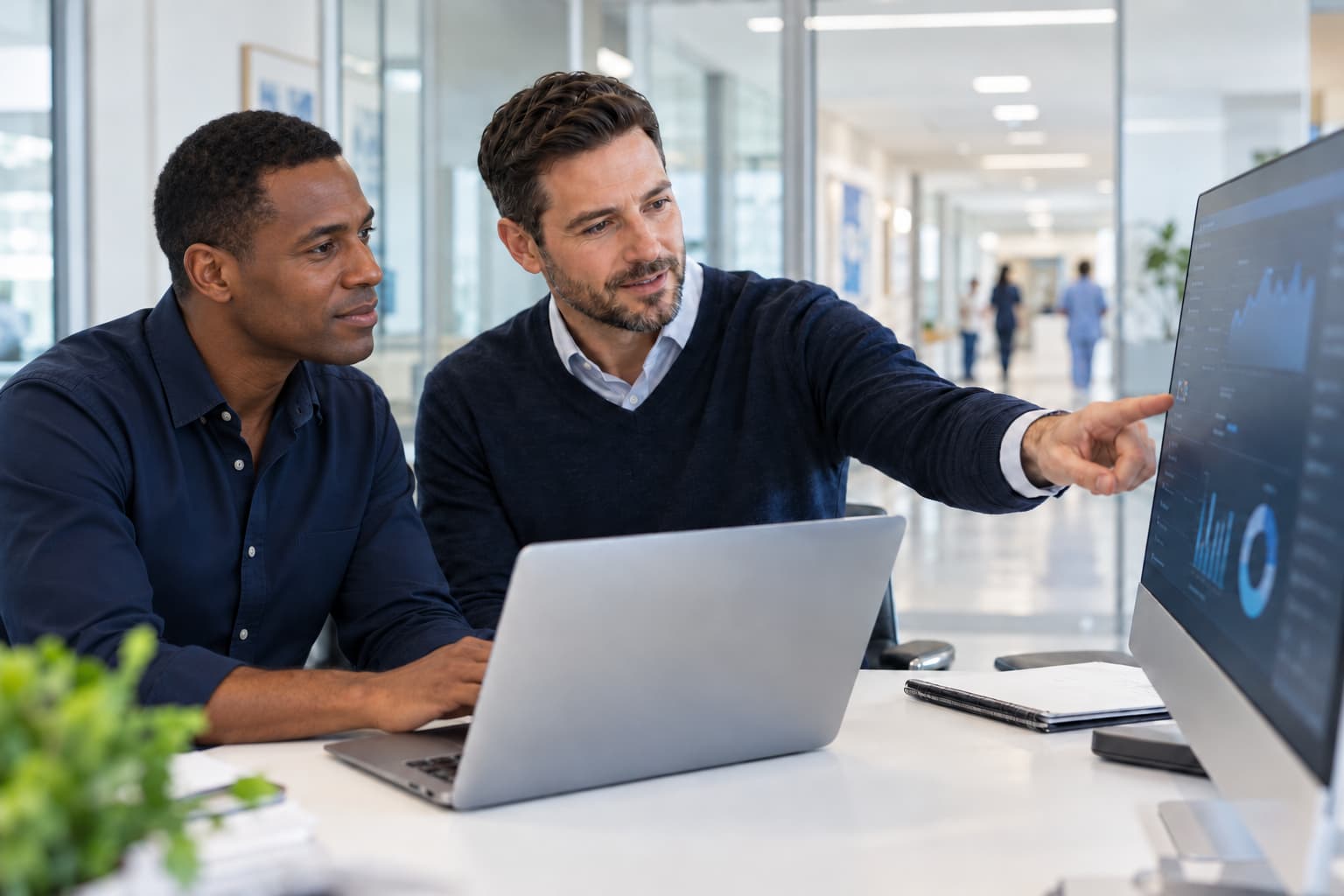 Two colleagues at a laptop in a bright hospital office, one pointing to charts on a large monitor