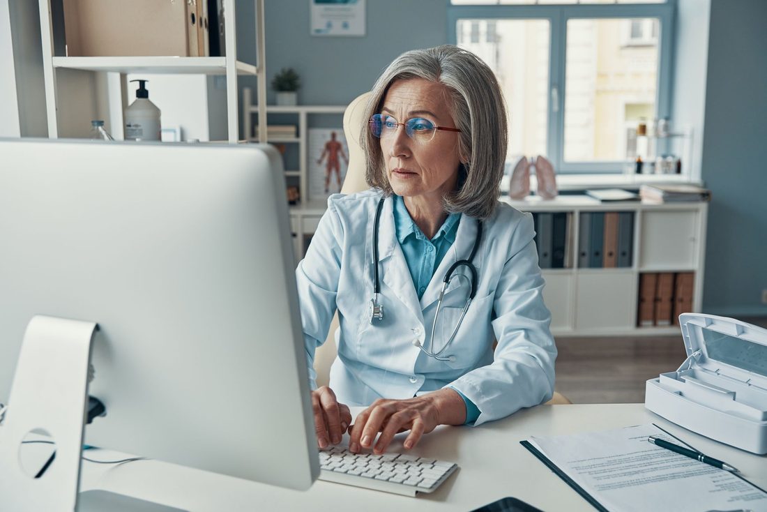 A doctor in a white coat working at a desktop computer in her consulting room